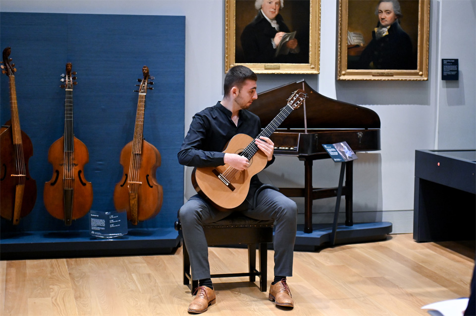 A male student playing the guitar, surrounded by musical instruments in a museum.